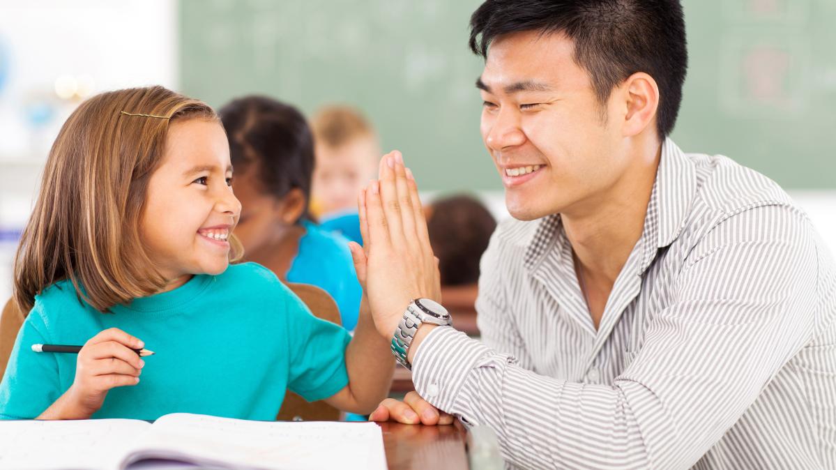 Young girl sitting at school desk giving high five to teacher