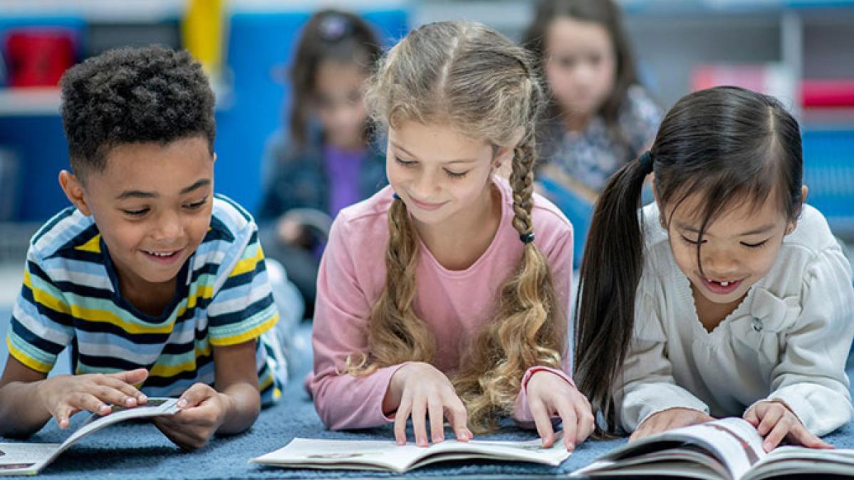 A young boy in a striped shirt, a young girl in a pink shirt and a young girl in a white shirt read books together. 