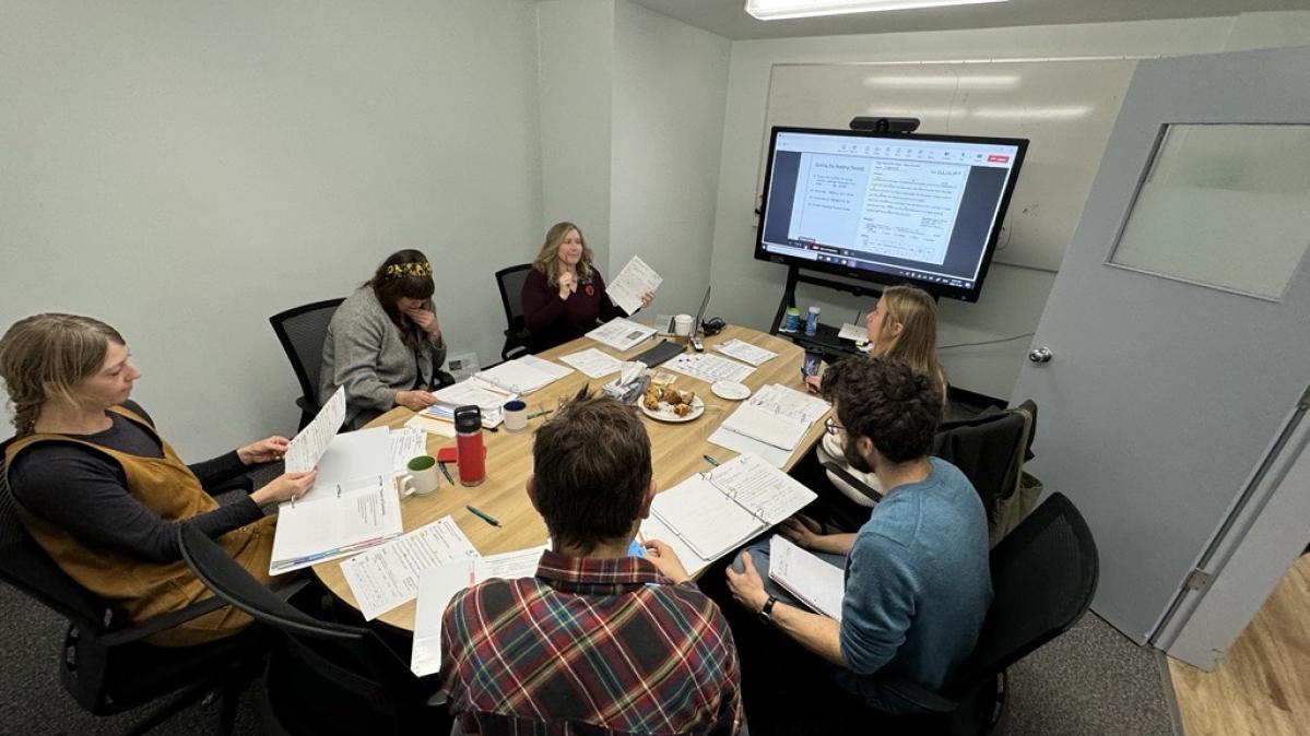 A group of people seated around a conference table facing a virtual screen. 