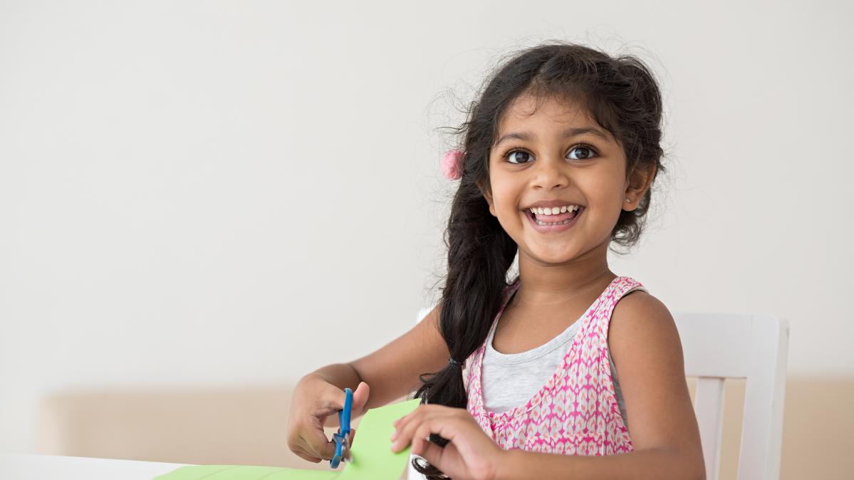 A preschool child with long dark hair smiling while they cut green paper with a pair of blue scissors.