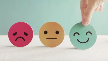 A red frowning face, a yellow neutral face and a green happy face cardboard cutouts in a row with a hand picking up the green one.