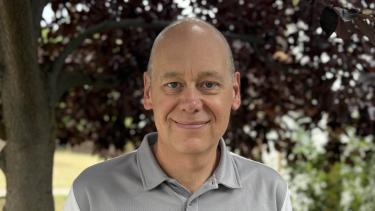 A man who is mostly bald wearing a grey and white short sleeved golf shirt standing and smiling in front of a leafy tree